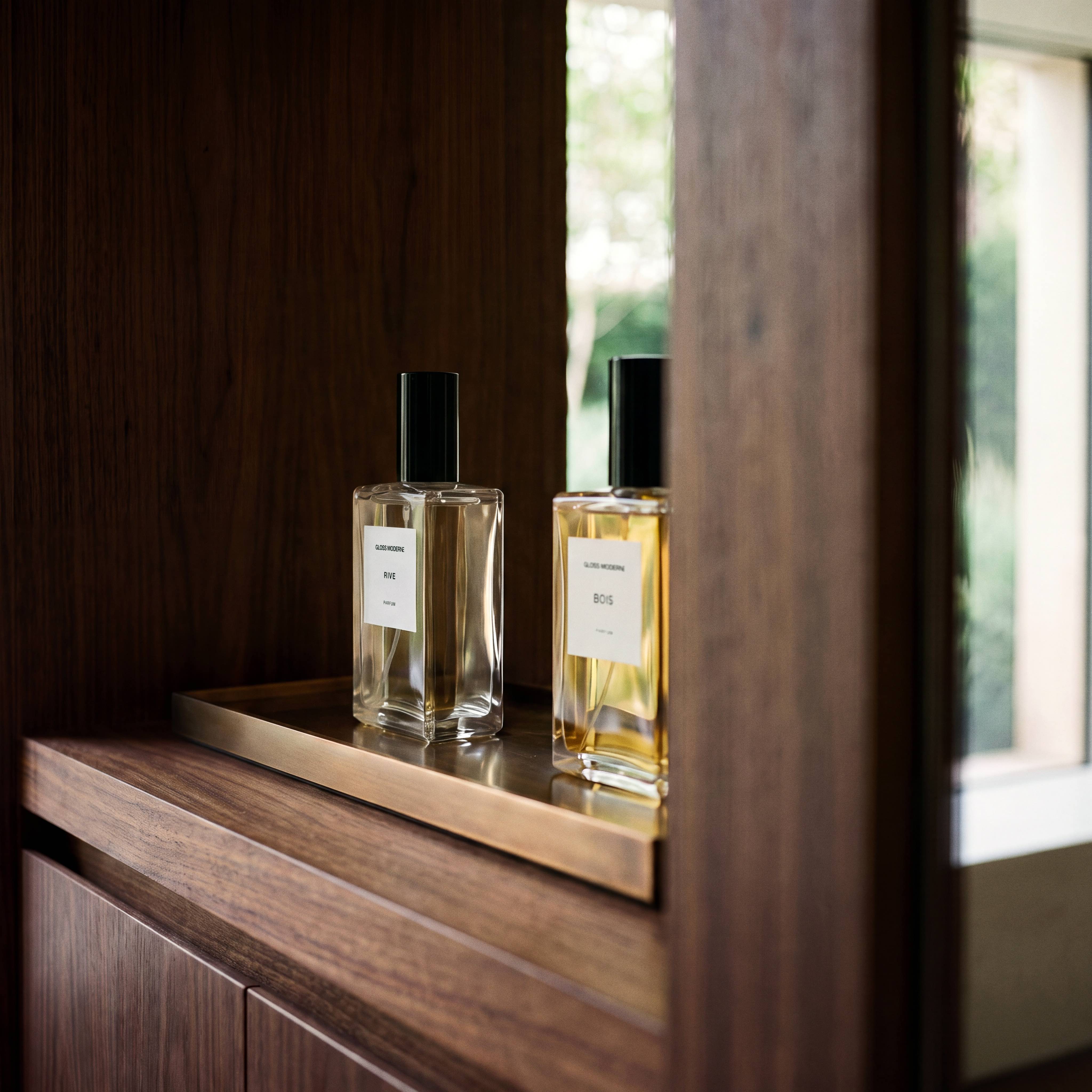 Two clear glass perfume bottles with black caps and white labels sit on a brass tray in a dark wood cabinet, with a window and greenery visible in the background.