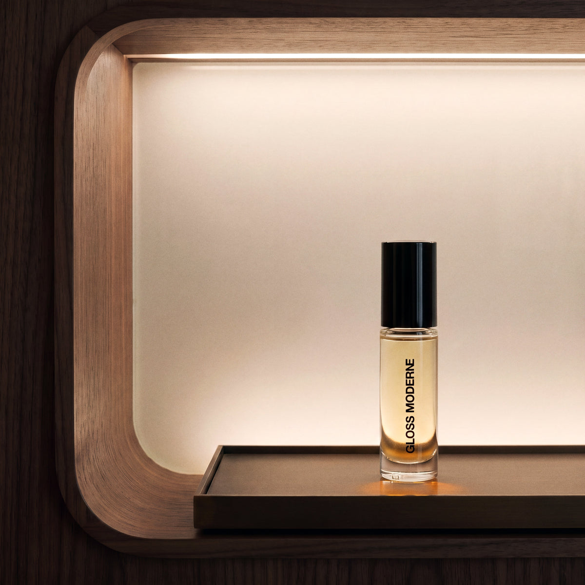 A clear glass perfume oil roll on bottle with a black cap and gold liquid, labeled 'GLOSS MODERNE', standing on a dark wooden shelf against a warm, backlit wood panel.