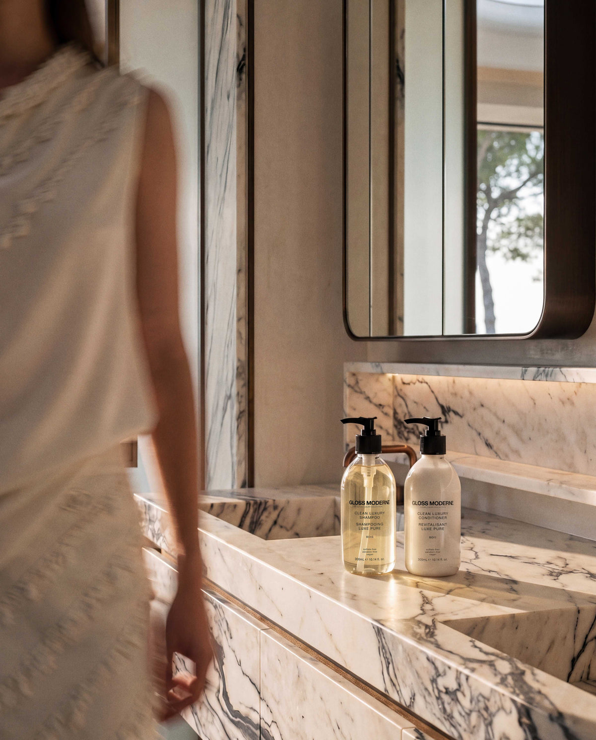 Two cosmetic bottles from the brand GLOSS MODERNE, one with a black pump and one with a clear pump, displayed on a white and gray marble vanity counter in a bathroom setting.