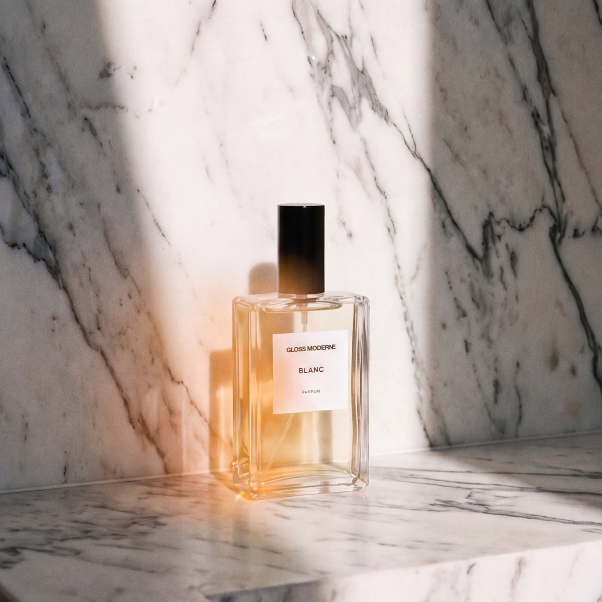 A clear glass perfume bottle with a black cap and a white label reading 'BLANC' sits on a white marble surface against a matching marble wall, lit by dramatic warm sunlight and shadow.