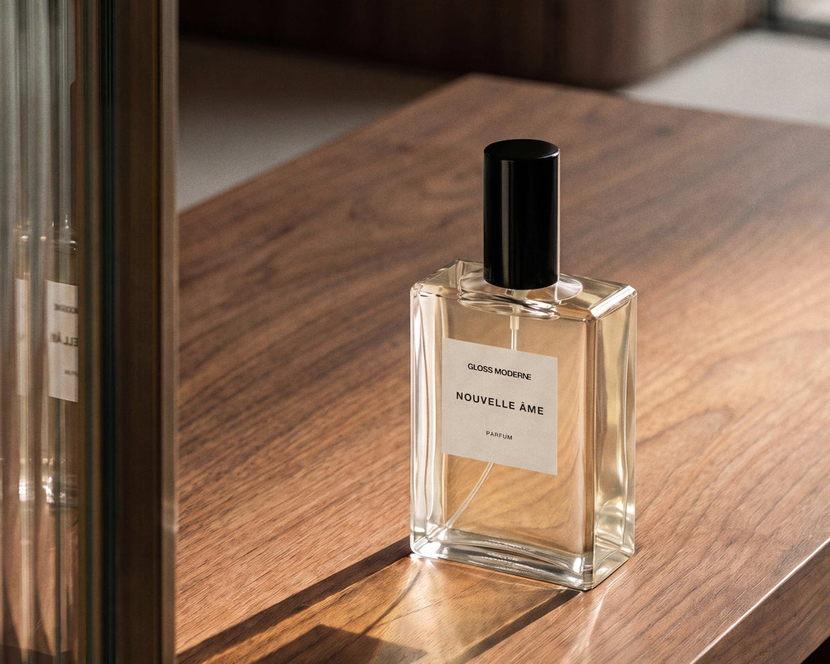 A clear glass perfume bottle with a black cap and a white label reading 'NOUVELLE ÀME' sits on a polished wooden table in a warm, modern interior setting.
